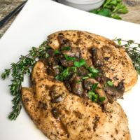 Seared chicken breasts with mushroom, onion, goat cheese sauce and a sprinkle of chopped parsley on a square white plate.