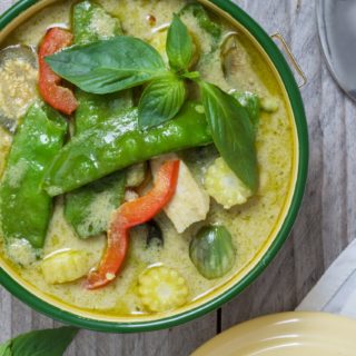 A ceramic bowl on a wooden table filled with green curry, red peppers, snow peas, chicken
