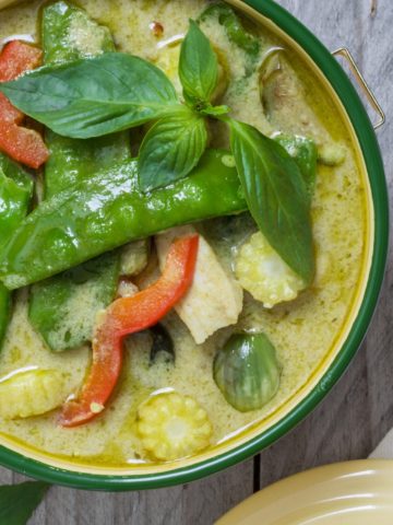 A ceramic bowl on a wooden table filled with green curry, red peppers, snow peas, chicken
