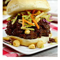 Thai seasoned ground beef on a toasted hamburger bun topped with broccoli slaw, on a white plate with peanuts scattered on it.