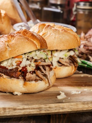 Closeup of two pulled pork sandwichses dressed with colewslaw and BBQ sauce on a wooden cutting board