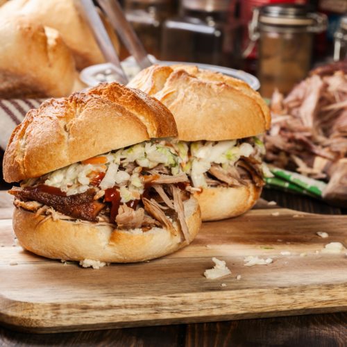 Closeup of two pulled pork sandwichses dressed with colewslaw and BBQ sauce on a wooden cutting board