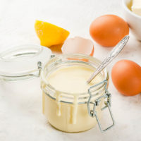 Hollandaise sauce in glass jar, with ingredients for cooking - eggs, butter, lemons. On a white concrete stone table.