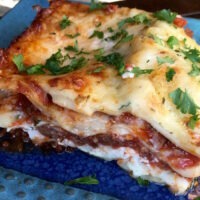 Close up of a slice of lasagna with layers of cheese, meat and noodles. Sprinkled with parsley. Served on a blue plate.