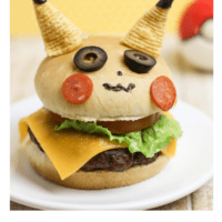 A Cheese burger on a bun decorated to look like Pikachu. 2 bugles are his ears, 2 slices of black olives are his eyes, two thin slices of pepperoni are his cheeks.
