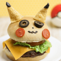 A Cheese burger on a bun decorated to look like Pikachu. 2 bugles are his ears, 2 slices of black olives are his eyes, two thin slices of pepperoni are his cheeks.