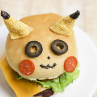 A Cheese burger on a bun decorated to look like Pikachu. 2 bugles are his ears, 2 slices of black olives are his eyes, two thin slices of pepperoni are his cheeks.
