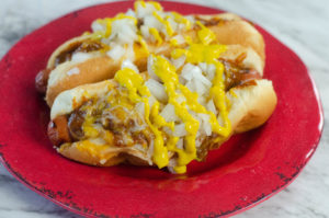 Hot dogs loaded with cheese and chili, topped with onion and mustard on a rustic red plate