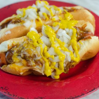 Hot dogs loaded with cheese and chili, topped with onion and mustard on a rustic red plate
