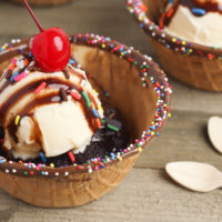 A waffle bowl rimmed with chocolate, and sprinkles, filled with a brownie, vanilla ice cream, fudge sauce, more sprinkles and a maraschino cherry