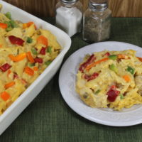 A white plate filled with Cheesy Chicken Noodle casserole, part of a white baking dish with the pasta in it.