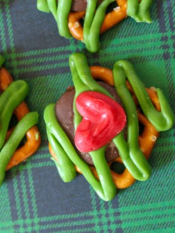 Square pretzels with a chocolate kiss melted on top and topped with green candy melt in a zig zag pattern and a red candy heart