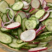 Thinkly sliced cucumbers, radish, red onion in a large glass mixing bowl. topped with toasted sesame seeds