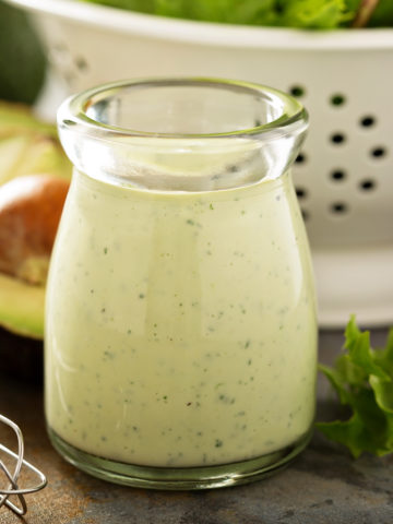 Homemade avocado ranch dressing in a small glass jar with fresh avocado cut in half and leafy greens in a colcander